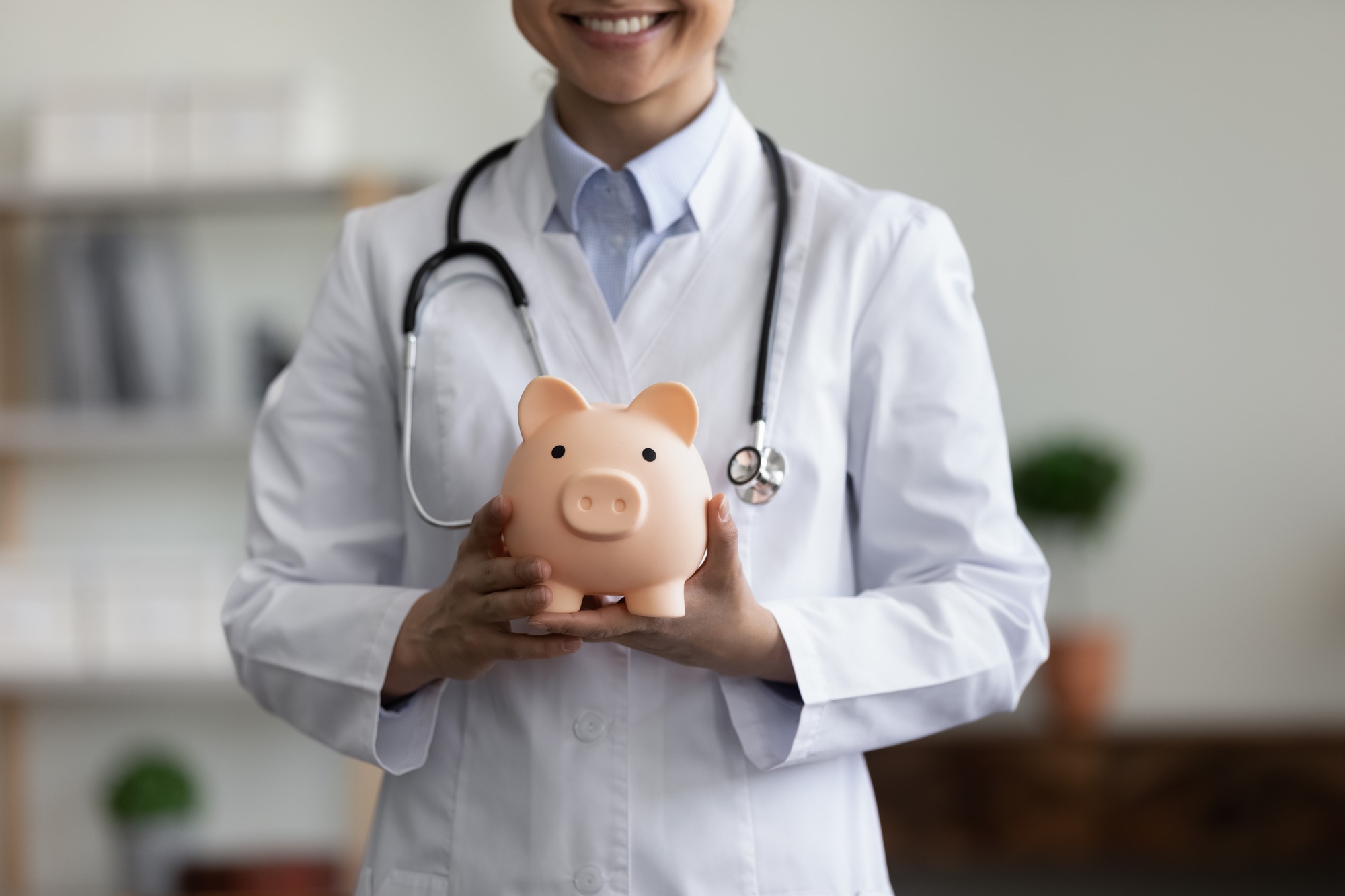 Close up happy young female GP doctor holding piggy bank in hands.