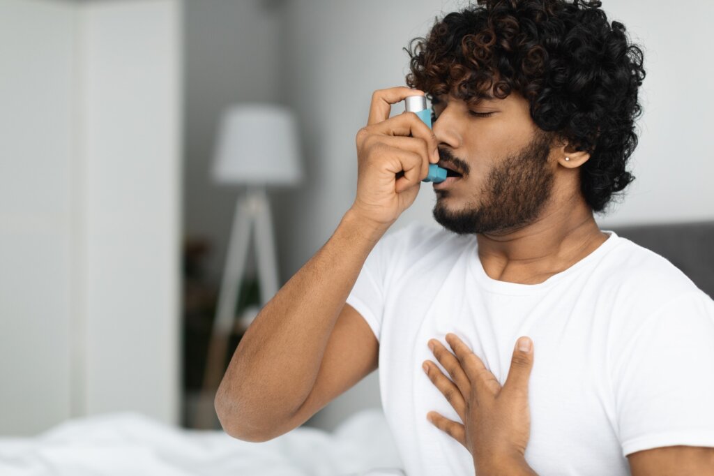 Indian guy suffering from asthma, using inhaler in bed, side view, copy space. Young hindu man woke up with asthma attack, touching chest and using nebulizer, bedroom interior