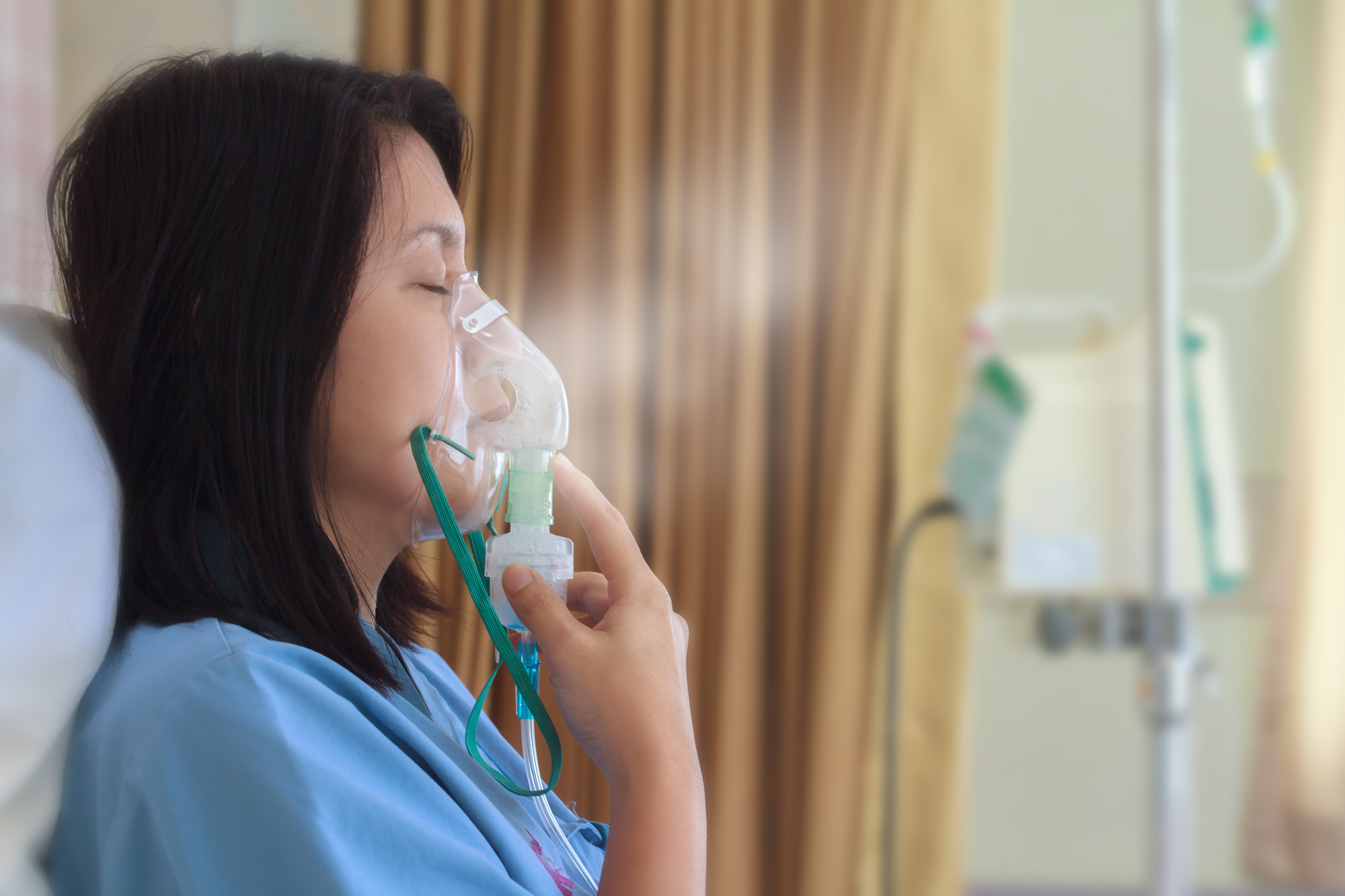 Sick beautiful female in blue cloth hold nasal mask with respiratory problem in hospital room. Asian woman patient inhalation therapy by the mask of inhaler with soft stream smoke from bronchodilator.