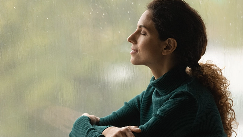 Music of rain. Side shot of serene latin woman relax by window with closed eyes listen sound of raindrops feel pleasure. Calm young lady enjoy breathing fresh ozonized air after rainstorm. Copy space.