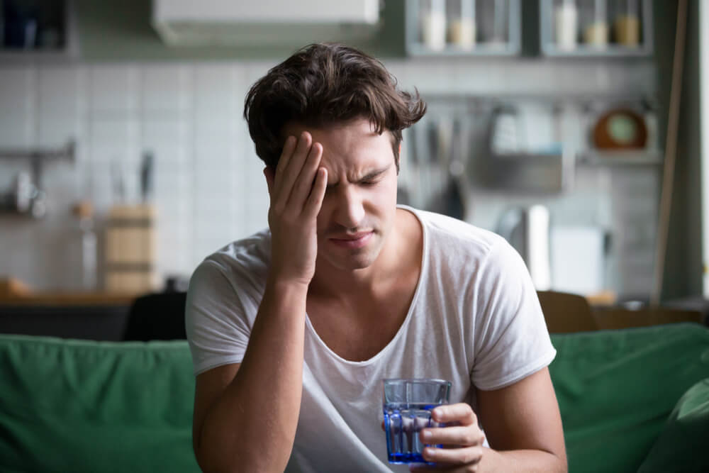Young man suffering from strong headache or migraine sitting with glass of water in the kitchen