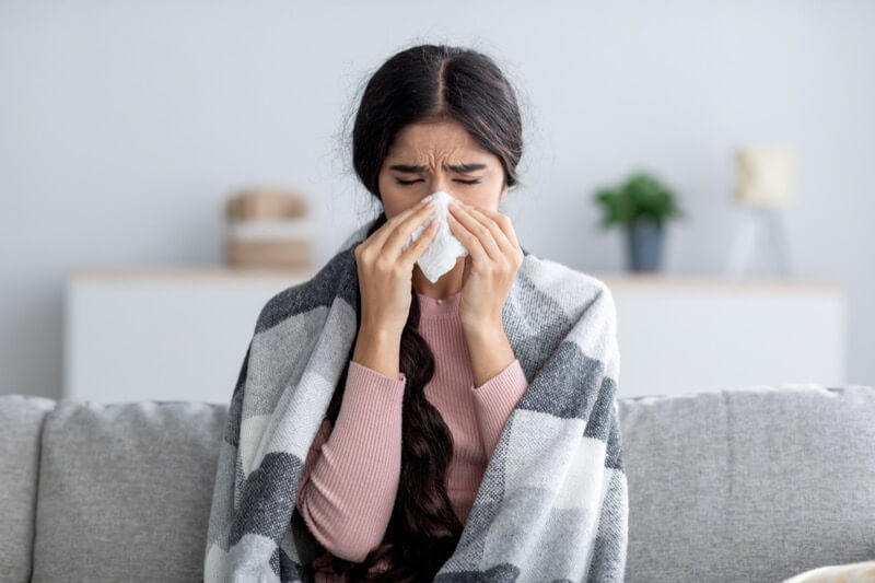 Unhappy sad young indian female in plaid suffering from fever and flu on sofa, blowing nose in napkin in living room interior. Covid-19 lockdown, treatment of illness, cold and runny, copy space