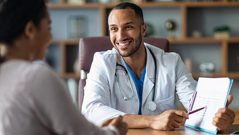 Cheerful young arabic general practitioner showing black lady patient treatment plan, african american woman attending therapist at newest private clinic, modern healthcare concept